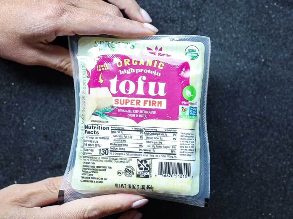 Two hands hold a package of organic high-protein super firm tofu. The packaging includes nutrition facts, a USDA organic label, and recycling symbols. The background is a textured black surface.